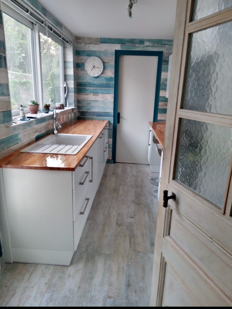 Cuisine rénovée avec meubles blancs, plan de travail en bois et évier sous une grande fenêtre.
